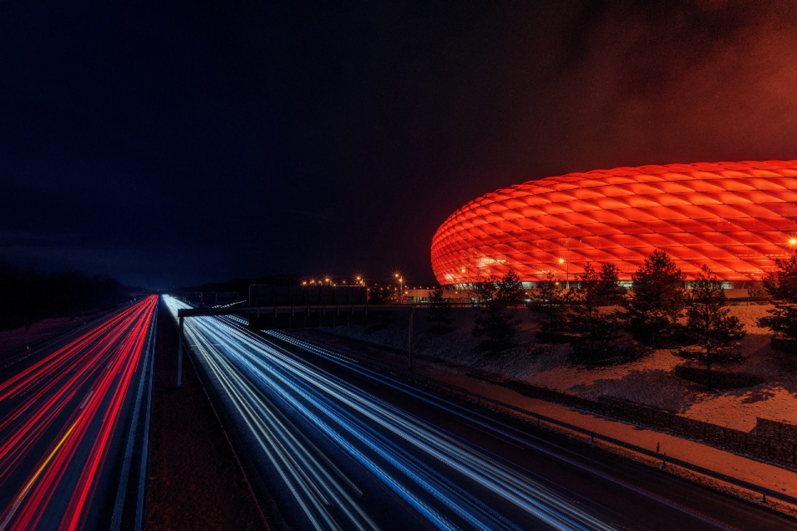 Stadium glowing red at night beside a highway of light trails